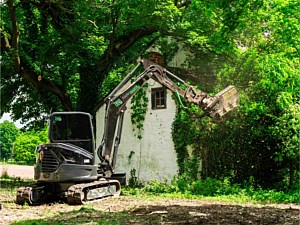Excavator with forestry mulcher working around abandoned farm house in green lane